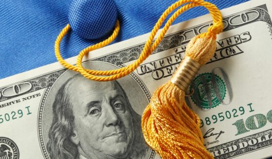 A stack of one hundred dollar bills on top of a blue graduation cap with a gold graduation tassel attached.